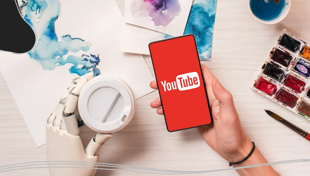 Designer at office desk with robot hand holding smartphone with YouTube app